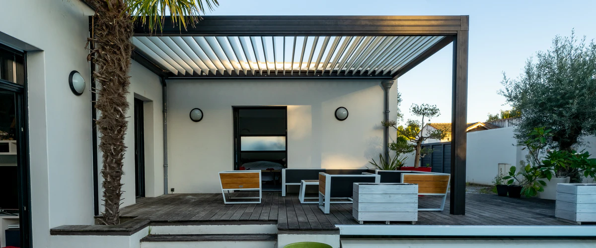 An attached modern black metal pergola with an adjustable louvered roof system installed over a wooden deck with minimalist outdoor furniture.