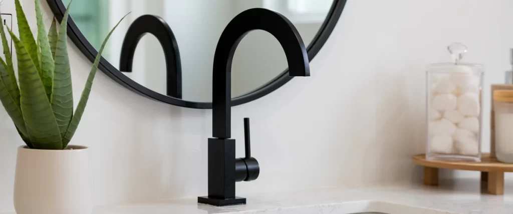 A minimalist matte black water fixture installed on a marble countertop next to a potted aloe vera plant and a round mirror.