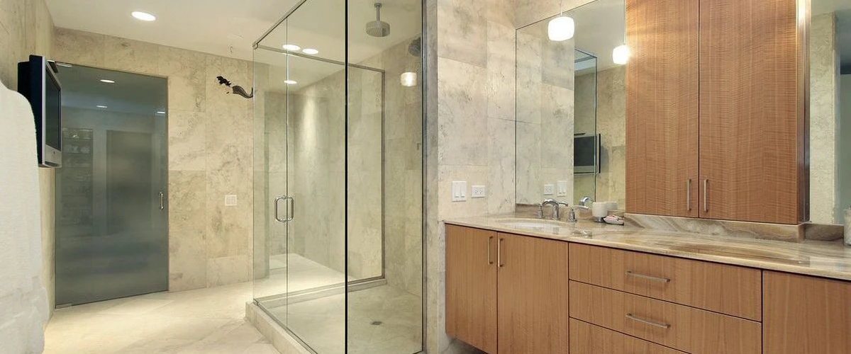 A spacious modern bathroom with tan stone tiled walls, a large frameless glass walk-in shower with dual showerheads, a frosted glass door, and a light wood double vanity.