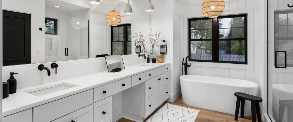 A bright modern white bathroom featuring a long double vanity with black hardware, shiplap walls, and woven pendant lights near a soaking tub.