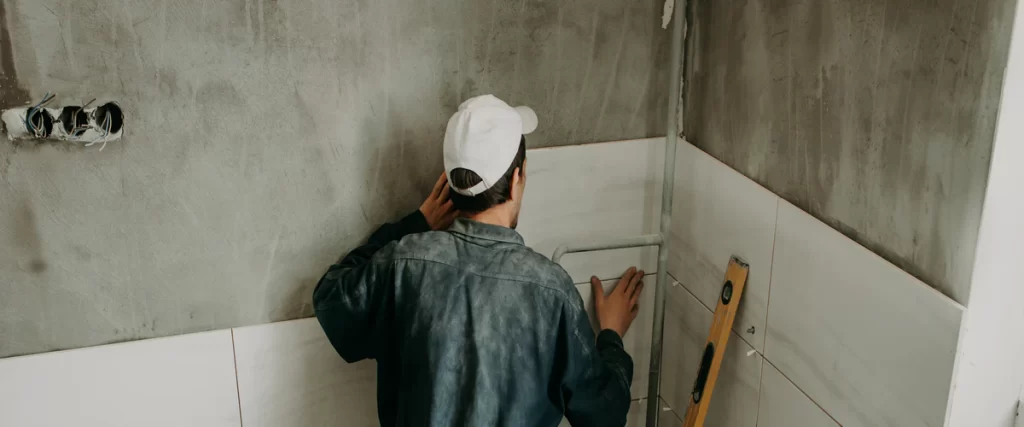A professional contractor in a white cap installing large white wall tiles over concrete during a residential bathroom renovation project.