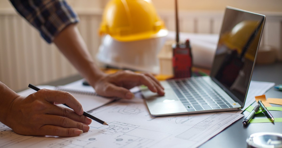A workspace featuring architectural blueprints, a laptop, a yellow hard hat, and a pencil, illustrating the preparation and questions to ask a contractor for a bathroom remodel.