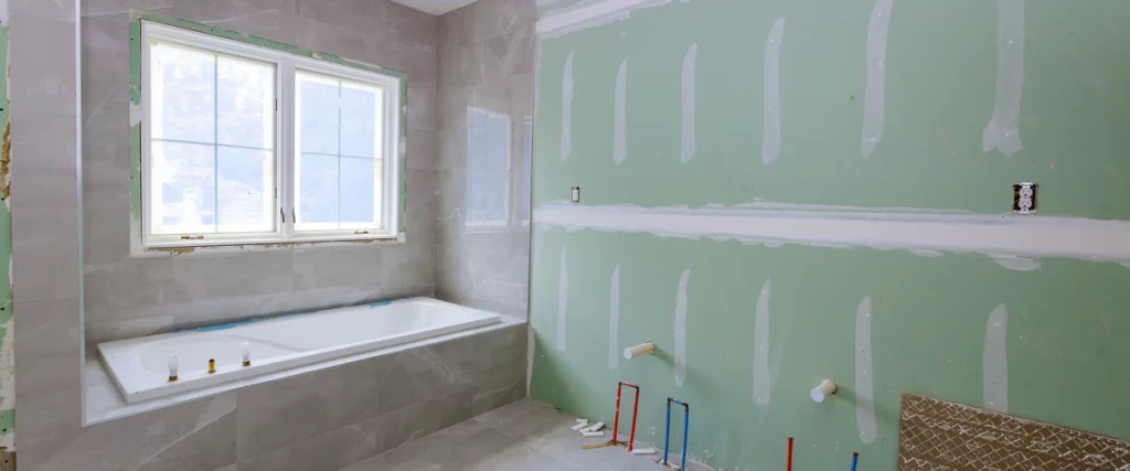 The interior construction phase of a bathroom remodel showing green moisture-resistant drywall, exposed plumbing pipes, and a grey tiled built-in tub.