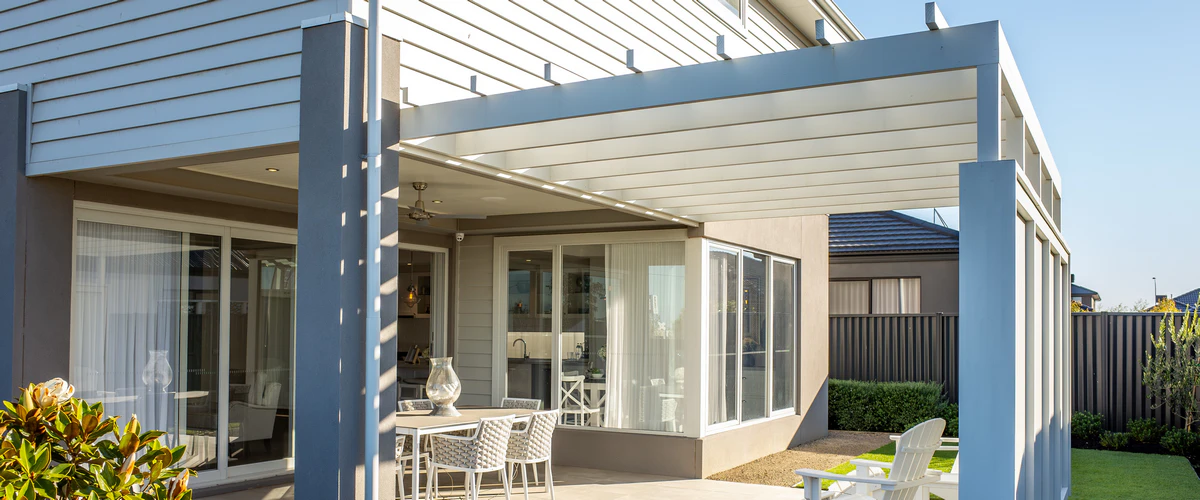 A large, custom white attached pergola featuring an integrated ceiling fan and lighting, covering a modern patio with white wicker dining furniture and glass doors.