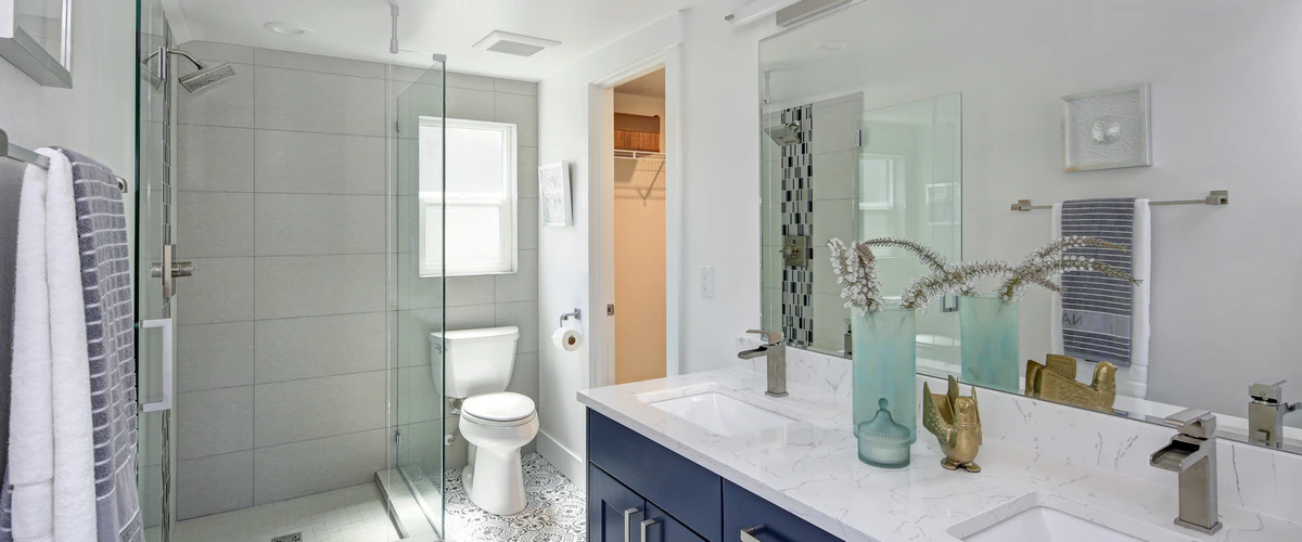 Bright white bathroom showcasing a dark blue vanity with white marble countertops, decorative patterned floor tiles, a glass shower enclosure, and modern fixtures.