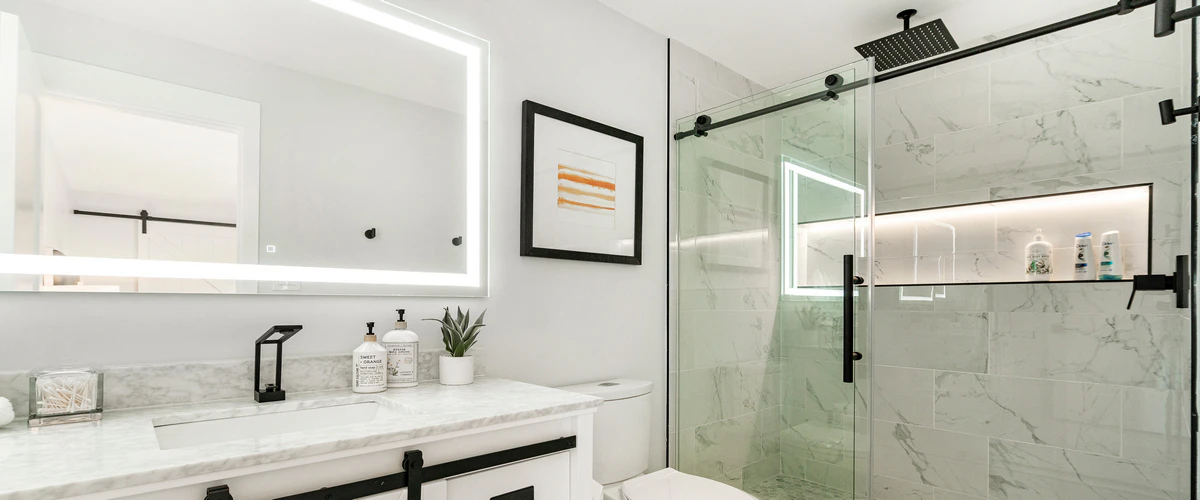 A luxury bathroom featuring a glass shower with black sliding hardware, a marble-topped white vanity, and a large rectangular LED-backlit mirror.