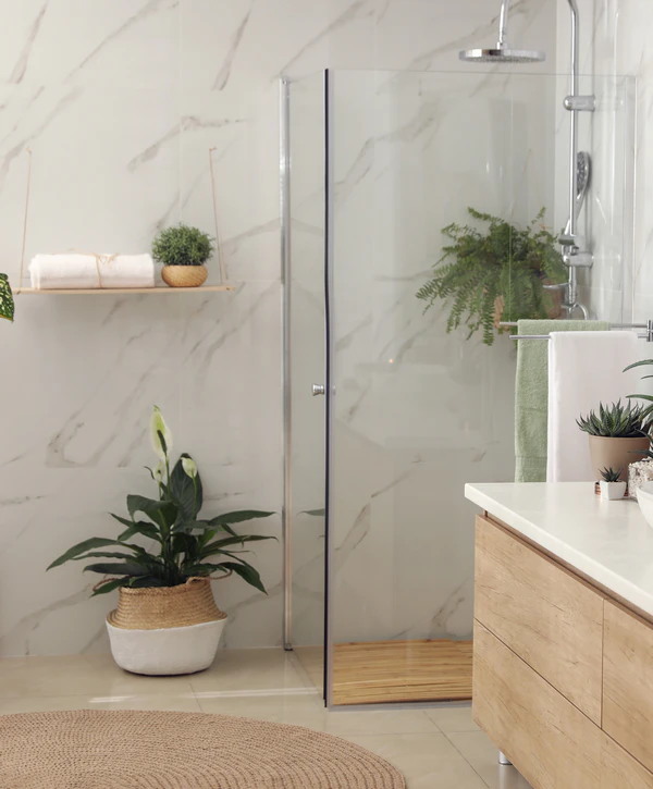 A modern bathroom shower remodeling project in Harker Heights, TX, featuring marble wall tiles, a glass enclosure with a wooden floor mat, and a light wood vanity accented by green houseplants.