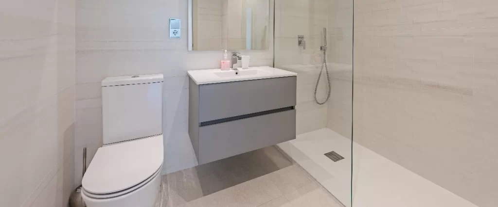 A well-designed small bathroom with light-colored floor tiles, featuring a toilet, a sleek grey floating vanity, and a space-saving glass shower enclosure.