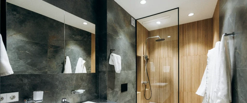 A contemporary dark stone bathroom highlighting a glass-enclosed shower with a wooden slat accent wall and built-in recessed storage for toiletries.