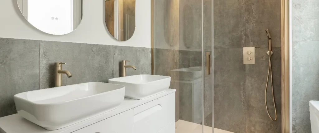 An elegant bathroom featuring a double vanity with vessel sinks, brushed gold faucets, and a frameless glass shower enclosure against a dark grey stone-textured wall.