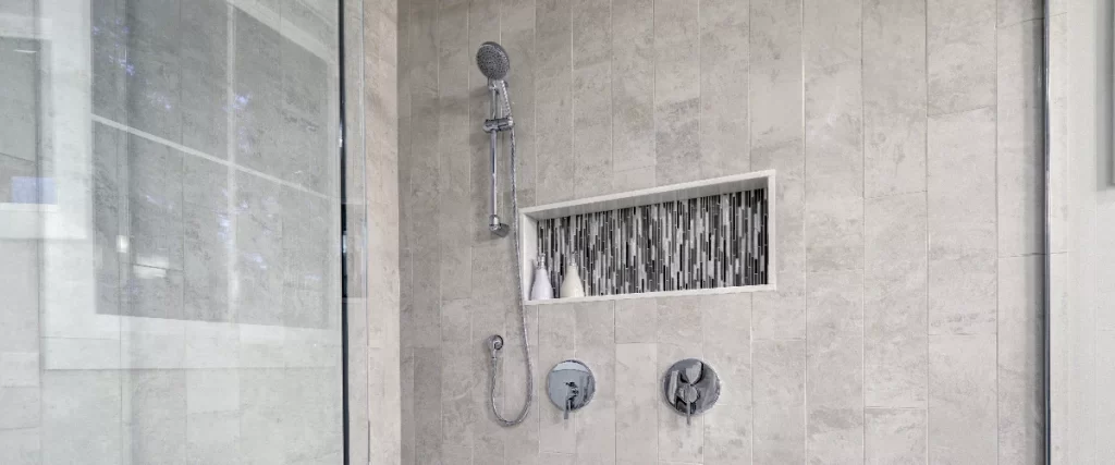 Close up of modern chrome shower fixtures mounted on light grey vertical tiles, featuring a built-in recessed niche with decorative mosaic tiles.