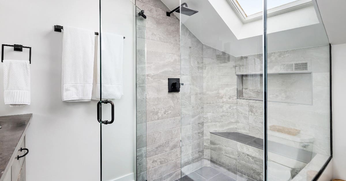 A modern bathroom showcasing walk in shower ideas with light grey textured tiles, a glass door, matte black fixtures, and a slanted ceiling with a skylight.