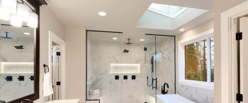 A bright and airy bathroom featuring a white marble-patterned glass shower illuminated by abundant natural light streaming in from a large ceiling skylight and a side window.